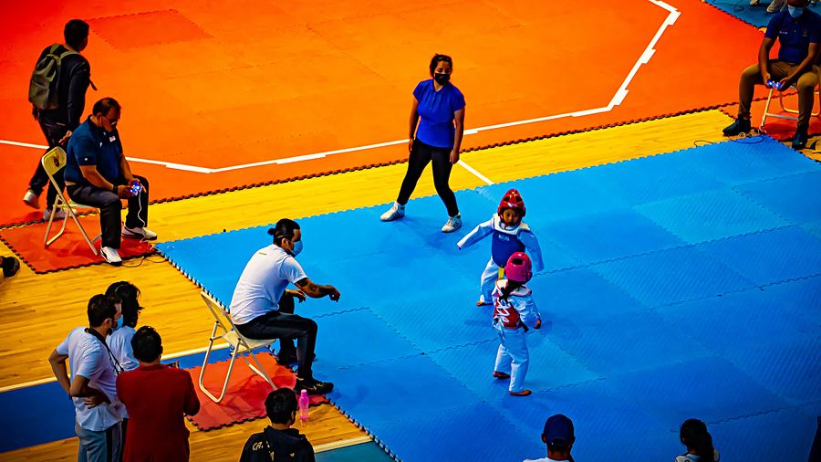 Dos niños pequeños con careta y peto compiten sobre un tatami azul; un entrenador los sigue desde la silla