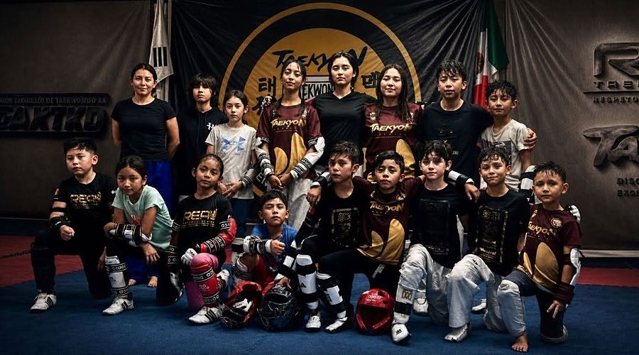 Alumnas y alumnos del club con playeras REAL y TaeKyon y protecciones, frente a la manta del dojang